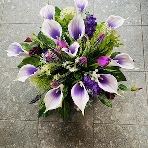 Peut inclure: Un arrangement floral composé de lys calla blancs et violets, de grappes de petites fleurs violet foncé et de divers feuillages verts. Le bouquet est disposé en forme arrondie avec de multiples tiges s'étendant vers l'extérieur, mettant en valeur un mélange de tons violets clairs et foncés sur des feuilles vertes vibrantes.