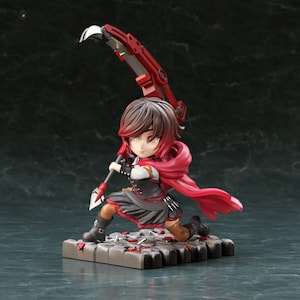 May include: A chibi-style figurine of a character with short dark red hair, wearing a black and red outfit with a flowing red cape. The character is kneeling on a stone-textured base and holding a large, mechanical red and silver scythe weapon.