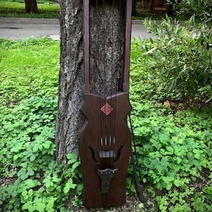 Może przedstawiać: Ciemnobrązowy drewniany instrument strunowy o wysokiej, prostokątnej ramie z zakrzywionymi górnymi rogami, czterema pionowymi strunami, centralną czerwoną geometryczną inkrustacją i podstawkiem. Pasujący ciemny drewniany smyczek opiera się o korpus instrumentu.