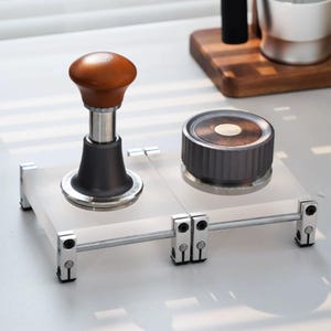 May include: A coffee tamper with a brown wooden handle and a black base, alongside a circular coffee distributor with a dark wood top and a metallic grey ribbed side, both resting on a clear acrylic stand with silver metal support rods.