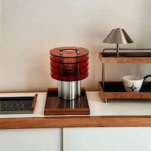 May include: A table lamp featuring a cylindrical silver metal base and five stacked translucent red circular acrylic discs. The lamp sits on a square wooden tray with a grid pattern. Beside it is a two-tiered wooden shelf with a metal lamp.