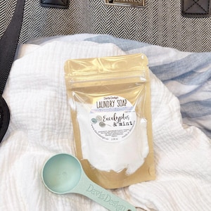 May include: A package of laundry soap with the words "Eucalyptus & Mint" and a light green measuring scoop. The soap is in a clear bag with a brown backing. The scoop is on a white and blue striped cloth.