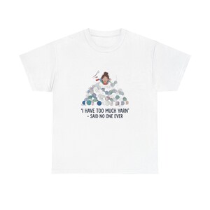 May include: A white short-sleeved t-shirt featuring a graphic of a person surrounded by a large pile of colourful yarn balls. Below the illustration, the text reads: 'I HAVE TOO MUCH YARN' - SAID NO ONE EVER. The design is centred on the chest area.