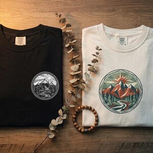 Pode incluir: Uma t-shirt preta e uma t-shirt branca, cada uma com um gráfico circular no peito. A t-shirt preta tem uma paisagem montanhosa monocromática, enquanto a branca exibe uma cena de montanha colorida com sol, árvores e um rio.