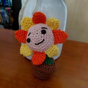 Peut inclure: Un jouet fleur au crochet avec un visage rose clair, des yeux en perles noires et un sourire brodé. La fleur possède des pétales alternés jaunes et orange et repose dans un petit pot marron au crochet avec un dessus vert représentant la terre.
