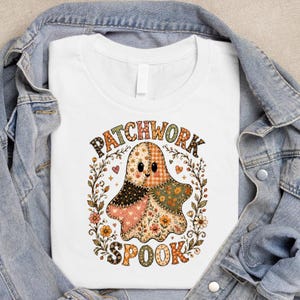 Peut inclure: Un t-shirt blanc avec un motif de fantôme composé de divers motifs de tissus aux couleurs automnales. Le fantôme est entouré d'une couronne florale et du texte PATCHWORK SPOOK dans une police décorative. Le t-shirt est sous une veste en jean bleu.