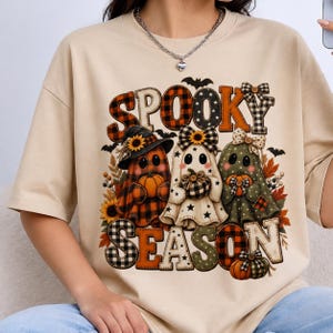 Puede incluir: Una camiseta beige de manga corta con un estampado gráfico que incluye el texto SPOOKY SEASON. El diseño muestra tres fantasmas con sombreros y lazos, rodeados de calabazas, girasoles, hojas de otoño y murciélagos en naranja, negro y blanco.
