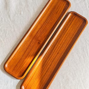 Op de afbeelding: Twee rechthoekige houten dienbladen met afgeronde hoeken en een warme, middelbruine houtnerfafwerking. De dienbladen zijn lang en smal, met een glad, licht verzonken oppervlak en opstaande randen die de natuurlijke houtstructuur tonen.