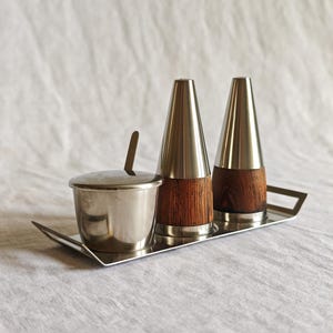 Puede incluir: Un juego de condimentos que incluye una bandeja de acero inoxidable con dos asas, un cuenco pequeño con tapa y cuchara, y dos saleros cónicos. Los saleros tienen tapas de acero inoxidable y bases de madera de color marrón oscuro.