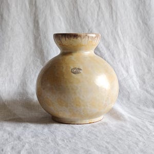 May include: A round ceramic vase with a wide, bulbous body and a narrow neck. The surface features a mottled cream and beige glaze with a darker brown drip pattern around the rim. A small, weathered silver sticker is centred on the front.
