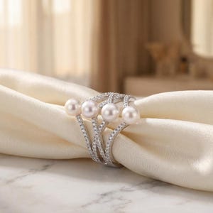 May include: A silver-toned ring featuring four round white pearls arranged in a row, set against a crisscrossing band encrusted with small clear stones. The ring is positioned around a folded piece of cream-colored fabric.
