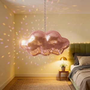 Può includere: Una lampada a sospensione a forma di nuvola ricoperta di piccole tessere a specchio riflettenti color oro rosa. L'apparecchio è appeso a una catena di metallo argentato e proietta riflessi di luce colorati sulle pareti e sul soffitto.