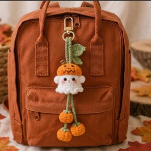 Op de afbeelding: Een roestkleurige canvas rugzak met een gehaakte sleutelhanger. De hanger toont een wit spookfiguurtje met een oranje pompoenhoed, hangend aan een groen koord met drie kleine oranje pompoenbedels en een groen blaadje.