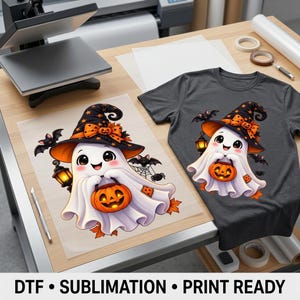 May include: A dark gray t-shirt featuring a graphic of a white ghost wearing a black witch hat with an orange bow, holding a jack-o'-lantern. The ghost is surrounded by small bats, a lantern, and a spider web. A matching transfer sheet is shown.