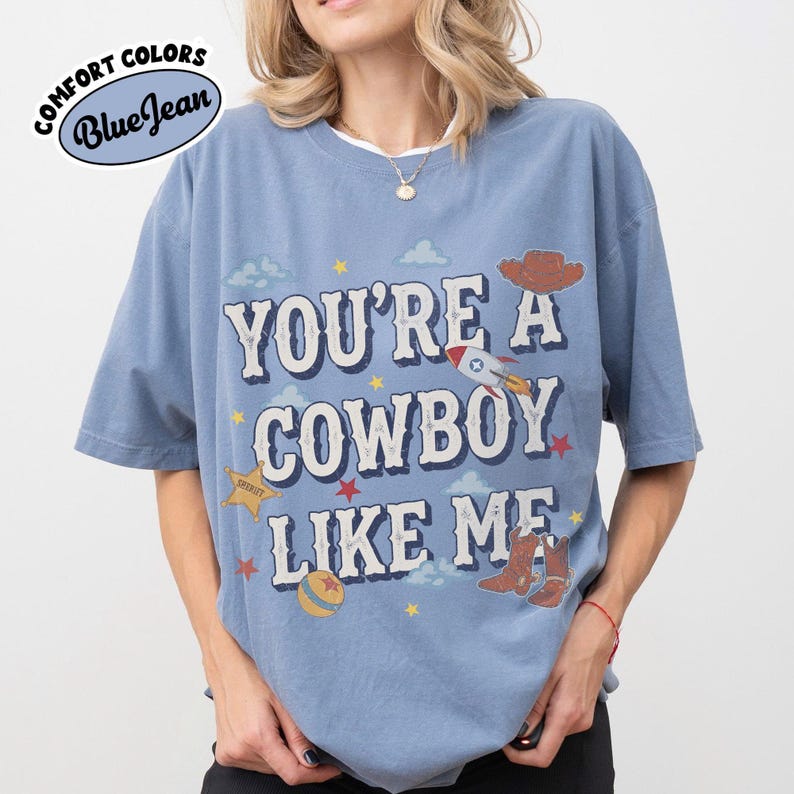 Op de afbeelding: Een blauw t-shirt met korte mouwen en de tekst "YOU'RE A COWBOY LIKE ME" in witte letters. De afbeelding bevat een bruine cowboyhoed, een raket, een gele sheriffster, een gele bal en bruine cowboylaarzen met rode sterren.