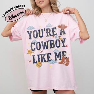 Op de afbeelding: Een lichtroze T-shirt met korte mouwen en de donkerblauwe tekst "YOU'RE A COWBOY LIKE ME" op de borst. De afbeelding bevat illustraties van een sheriff-badge, een raket, een cowboyhoed, bruine laarzen, een bal, wolken en sterren.