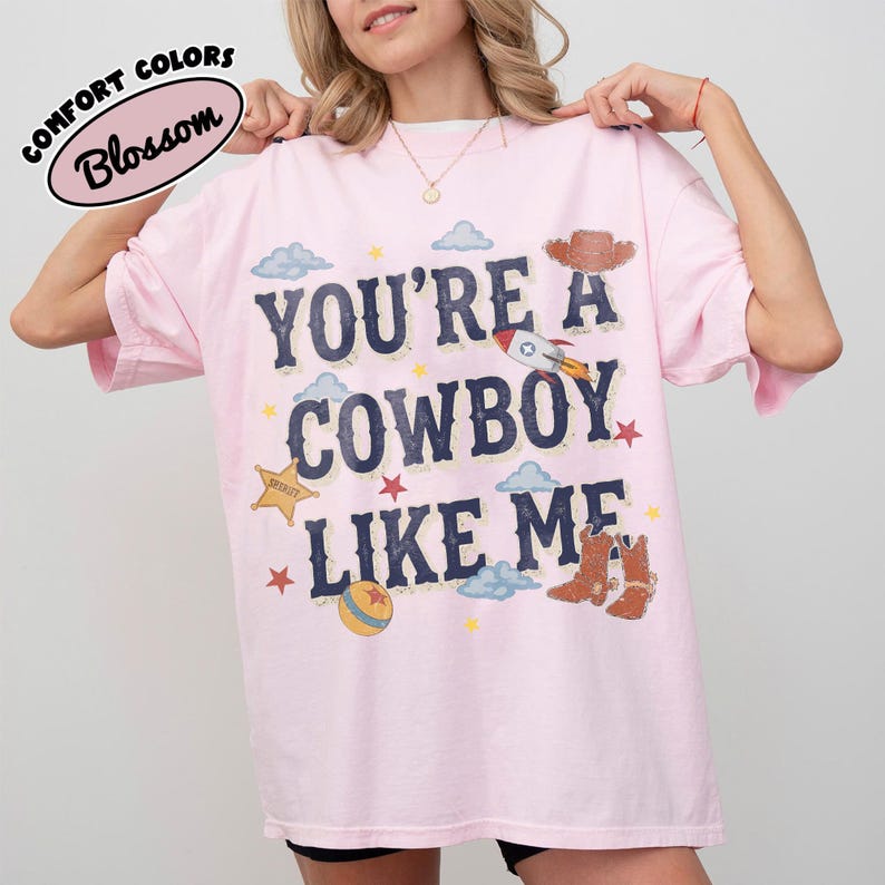 Op de afbeelding: Een lichtroze T-shirt met korte mouwen en de donkerblauwe tekst "YOU'RE A COWBOY LIKE ME" op de borst. De afbeelding bevat illustraties van een sheriff-badge, een raket, een cowboyhoed, bruine laarzen, een bal, wolken en sterren.