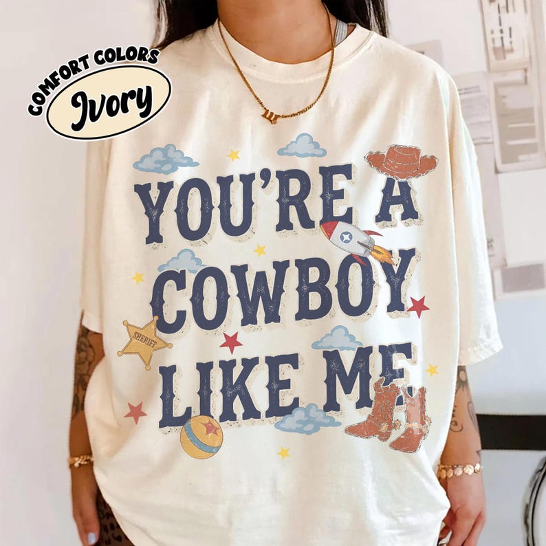 Op de afbeelding: Een ivoorkleurig t-shirt met de donkerblauwe tekst "YOU'RE A COWBOY LIKE ME" en grafische illustraties van een cowboyhoed, een raket, een sheriffster, een bal en cowboylaarzen, geaccentueerd door kleine sterren en lichtblauwe wolken.