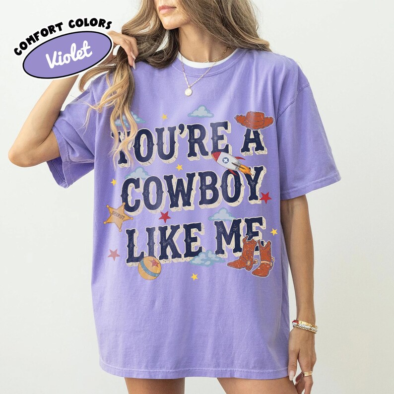 Op de afbeelding: Een violetkleurig T-shirt met korte mouwen en de tekst "YOU'RE A COWBOY LIKE ME" in donkerblauwe letters. De afbeelding bevat een bruine cowboyhoed, een raket, een sheriffster, een bal en bruine cowboylaarzen, geaccentueerd door kleine sterren en wolken.