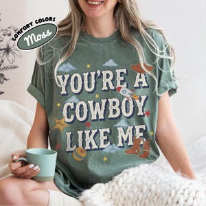 Op de afbeelding: Een mosgroen t-shirt met korte mouwen en de tekst "YOU'RE A COWBOY LIKE ME" in een verweerd lettertype. De afbeelding bevat illustraties van een raket, een sheriffster, een bal, wolken, sterren en cowboylaarzen in diverse kleuren.
