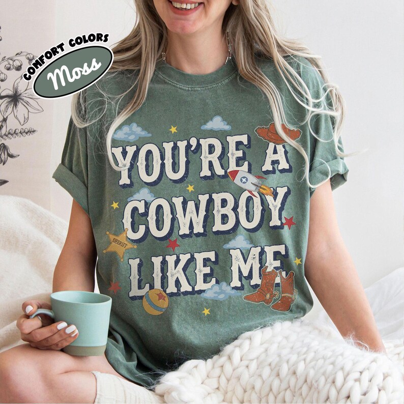 Op de afbeelding: Een mosgroen t-shirt met korte mouwen en de tekst "YOU'RE A COWBOY LIKE ME" in een verweerd lettertype. De afbeelding bevat illustraties van een raket, een sheriffster, een bal, wolken, sterren en cowboylaarzen in diverse kleuren.