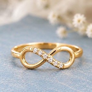 May include: A gold ring featuring an infinity symbol design on the front. The infinity loop is accented with five small, clear, round-cut stones embedded along one side of the metal band, creating a polished and reflective metallic finish.