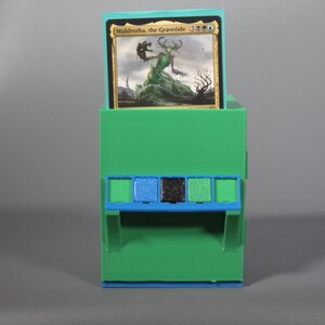 K&ouml;nnte beinhalten: Eine hellgr&uuml;ne Kunststoff-Sammelkartenbox mit einer blauen horizontalen Akzentleiste und drei kleinen quadratischen Eins&auml;tzen in Blau, Schwarz und Gr&uuml;n. Eine Magic: The Gathering-Karte mit dem Titel Muldrotha, the Gravetide steckt im oberen Schlitz.
