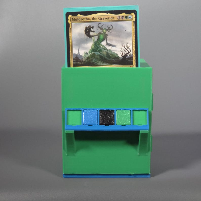 K&ouml;nnte beinhalten: Eine hellgr&uuml;ne Kunststoff-Sammelkartenbox mit einer blauen horizontalen Akzentleiste und drei kleinen quadratischen Eins&auml;tzen in Blau, Schwarz und Gr&uuml;n. Eine Magic: The Gathering-Karte mit dem Titel Muldrotha, the Gravetide steckt im oberen Schlitz.