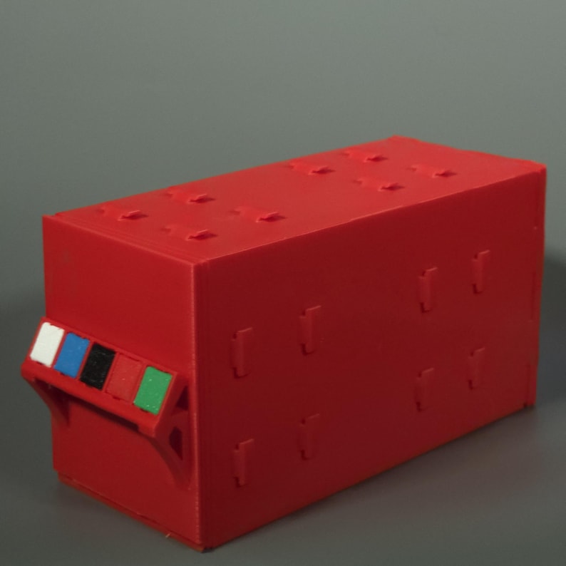 K&ouml;nnte beinhalten: Eine rechteckige rote Aufbewahrungsbox in modularem Design mit sichtbaren Verbindungsstiften an den Seiten und auf der Oberseite. Ein Griff an der Vorderseite zeigt f&uuml;nf kleine quadratische Eins&auml;tze in Wei&szlig;, Blau, Schwarz, Rot und Gr&uuml;n.