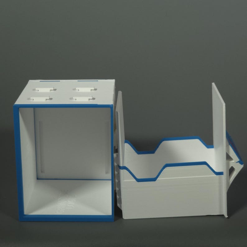 K&ouml;nnte beinhalten: Ein modularer Aufbewahrungsorganizer in Wei&szlig; und Blau, bestehend aus einer rechteckigen Box mit hohlem Innenraum und einer separaten, oben offenen Ablage mit eingekerbten Seitenw&auml;nden. Die Teile bestehen aus starren Platten mit blauem Rand.