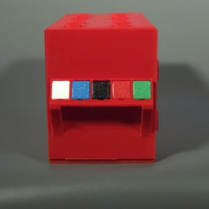 K&ouml;nnte beinhalten: Ein rechteckiger roter Kunststoffblock mit einer Reihe von f&uuml;nf kleinen quadratischen Tasten in Wei&szlig;, Blau, Schwarz, Rot und Gr&uuml;n. Die Oberseite des roten Blocks weist ein Gitter aus kreisf&ouml;rmigen Noppen auf und die Vorderseite enth&auml;lt eine eingelassene horizontale Ablage.