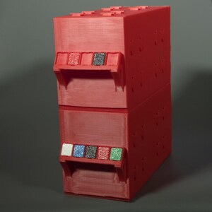 K&ouml;nnte beinhalten: Eine rote, rechteckige, 3D-gedruckte Aufbewahrungseinheit mit zwei &uuml;bereinanderliegenden F&auml;chern. Der obere Teil verf&uuml;gt &uuml;ber zwei kleine quadratische Eins&auml;tze in Rot und Schwarz. Der untere Teil hat f&uuml;nf kleine quadratische Eins&auml;tze in Wei&szlig;, Blau, Schwarz, Rot und Gr&uuml;n.