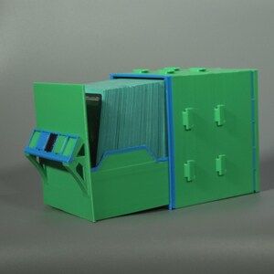 K&ouml;nnte beinhalten: Eine gr&uuml;ne Kunststoff-Aufbewahrungsbox mit blauen Akzenten und einer ausziehbaren Schublade, die mit einem Stapel gr&uuml;ner Karten gef&uuml;llt ist. Das Geh&auml;use hat ein modulares Design mit hervorstehenden rechteckigen Laschen zum Stapeln.