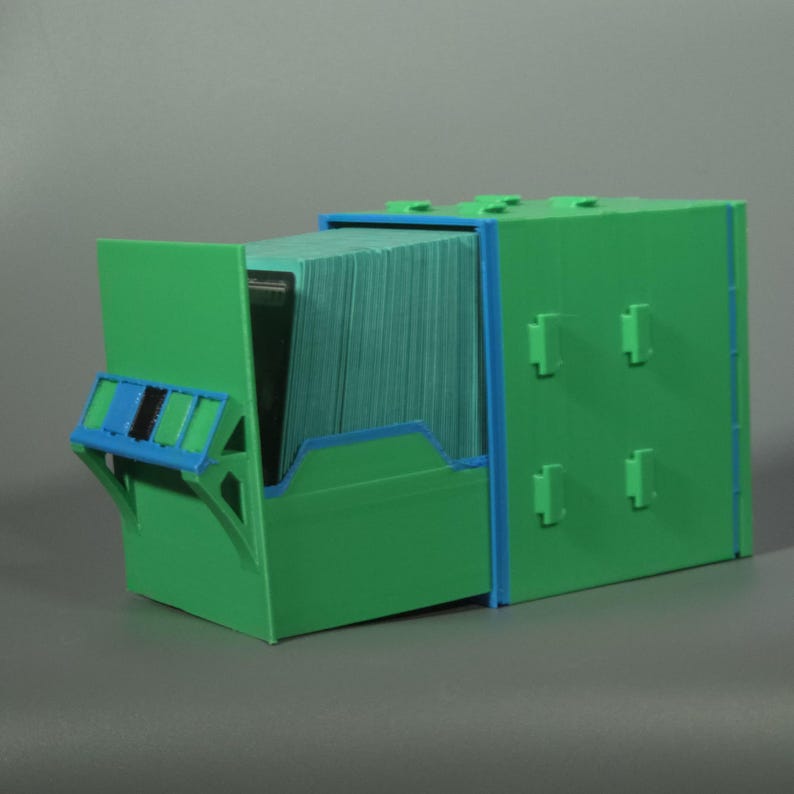 K&ouml;nnte beinhalten: Eine gr&uuml;ne Kunststoff-Aufbewahrungsbox mit blauen Akzenten und einer ausziehbaren Schublade, die mit einem Stapel gr&uuml;ner Karten gef&uuml;llt ist. Das Geh&auml;use hat ein modulares Design mit hervorstehenden rechteckigen Laschen zum Stapeln.