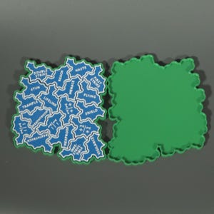 May include: Two irregular, cloud-shaped game tokens. One token is solid green. The other is blue with white outlines and text, featuring game keywords: Double Strike, Haste, First Strike, Flying, Shield, Vigilance, Indestructible, Hexproof, Menace, Reach, Stun, Trample, Lifelink, Deathtouch, and symbols +1/+1 and -1/-1.