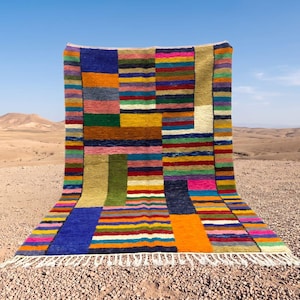 Peut inclure: Un tapis rectangulaire présentant un motif géométrique multicolore et vibrant composé de rayures horizontales et verticales dans des tons de bleu, orange, vert, jaune, rose et rouge, terminé par une frange nouée blanche sur le bord inférieur.