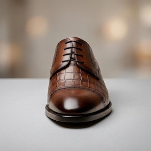 May include: A brown leather oxford shoe featuring a textured crocodile-embossed pattern on the upper, a smooth rounded toe cap with decorative brogue detailing along the seam, and dark brown laces threaded through five pairs of eyelets.