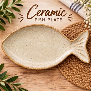 May include: A fish-shaped ceramic plate with a speckled off-white glaze and a brown rim. The plate features a textured surface and a defined tail fin design. The text "Ceramic FISH PLATE" is printed in brown above the plate in the image.