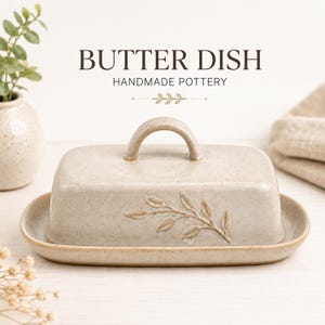 Peut inclure: Un beurrier rectangulaire en céramique beige mouchetée avec un couvercle assorti doté d'une poignée incurvée et d'un motif de branche de feuilles en relief. Le texte "BUTTER DISH HANDMADE POTTERY" est imprimé au-dessus en brun foncé.
