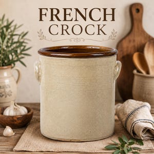 May include: A cylindrical beige ceramic crock with a dark brown glazed rim and two small side handles. The text FRENCH CROCK is printed in dark brown above the vessel. The crock has a smooth, matte finish and a wide, open top opening.