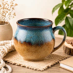 May include: A round ceramic mug featuring a two-tone glaze with a deep blue upper section that transitions into a speckled tan and brown base. The mug has a wide, bulbous body, a smooth rim, and a curved handle on the right side of the vessel.