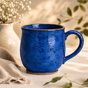 May include: A round ceramic mug with a vibrant cobalt blue glaze featuring a speckled texture and a dark brown rim. The mug has a curved body, a sturdy handle on the right side, and a visible unglazed brown base at the bottom of the vessel.