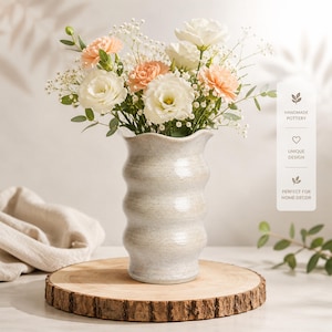 May include: A light beige, speckled ceramic vase with a wavy, tiered silhouette and a scalloped rim. The vase is filled with a bouquet of white and peach-colored carnations, white eustoma flowers, and delicate baby's breath with green eucalyptus leaves.