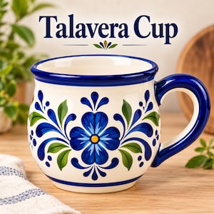 Puede incluir: Una taza de cerámica con cuerpo redondeado, base de color crema decorada con motivos florales azules y hojas verdes pintados a mano. El borde y el asa son de color azul sólido. El texto Talavera Cup aparece impreso sobre la taza en azul.