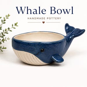 May include: A blue and cream ceramic bowl shaped like a whale, featuring a glossy dark blue exterior, a cream-coloured underside with vertical grooves, a tail fin, and a small side fin. The text above reads Whale Bowl HANDMADE POTTERY.
