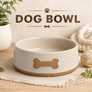 Puede incluir: Un cuenco redondo de cerámica de color blanco roto moteado con un emblema en relieve en forma de hueso marrón en la parte frontal. El texto DOG BOWL aparece impreso sobre el cuenco, junto a una pequeña huella y un icono de hueso.
