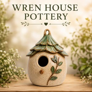 May include: A ceramic birdhouse with a speckled beige body and a layered green roof. The front features a circular entrance hole, a small brown knob, and a raised green leaf branch design. The text WREN HOUSE POTTERY is displayed above the object.