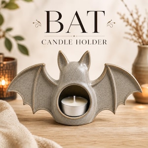 Puede incluir: Un portavelas de cerámica gris con forma de murciélago, acabado moteado y una abertura circular central que contiene una vela de té blanca. El texto BAT CANDLE HOLDER aparece encima en una fuente serif de color marrón oscuro con adornos.