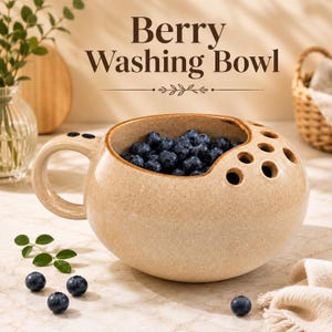 May include: A beige ceramic berry washing bowl with a rounded shape, a side handle, and a series of drainage holes on the top rim. The bowl is filled with fresh blueberries, and the text Berry Washing Bowl is displayed above the object.