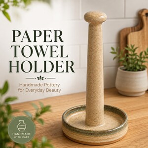 Pode incluir: Um suporte de papel toalha de cerâmica bege salpicada com um poste central alto e uma base circular com uma borda verde escura. O texto PAPER TOWEL HOLDER e Handmade Pottery for Everyday Beauty são visíveis, junto com um logo HANDMADE WITH CARE.